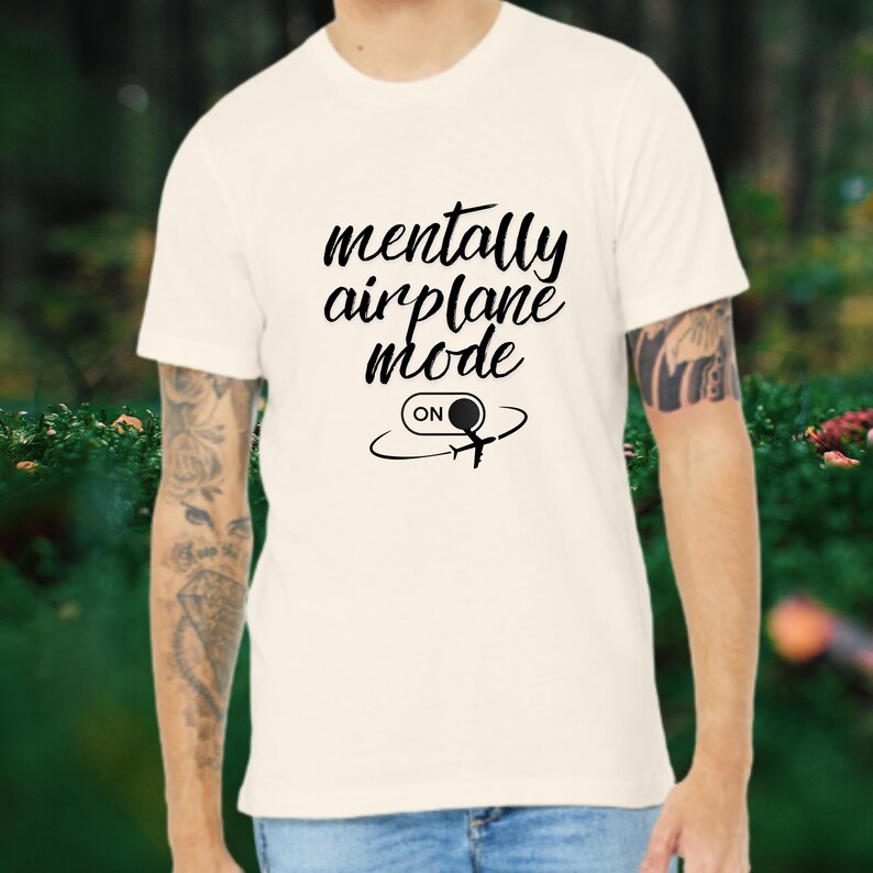 Może przedstawiać: Kremowa koszulka z napisem "mentally airplane mode" w czarnym skrypcie. Poniżej czarna grafika samolotu z zakrzywionymi liniami i słowem "ON" w okręgu.