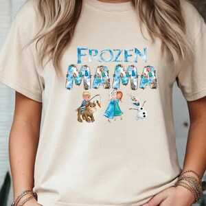 Pu&ograve; includere: T-shirt beige con la scritta "FROZEN MAMA" in lettere blu e bianche. La parola "FROZEN" &egrave; in un carattere blu ghiaccio. La parola "MAMA" &egrave; in un carattere blu e bianco con personaggi dei cartoni animati del film Frozen.