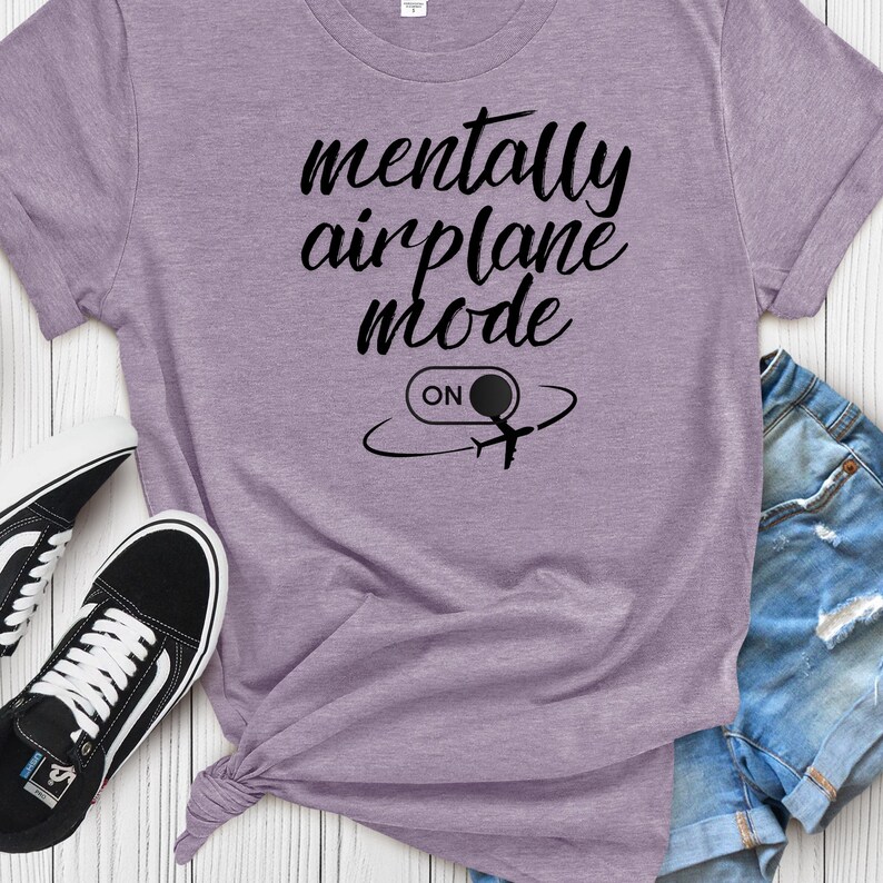 Może przedstawiać: Jasnofioletowa koszulka z czarnym napisem "mentally airplane mode". Poniżej tekstu znajduje się grafika samolotu ze słowem "ON" w okręgu. Widoczne są czarno-białe trampki i dżinsowe szorty.