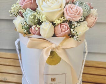 Può includere: Una composizione floreale di candele a forma di rosa in una scatola rotonda bianca. Le candele sono nei toni del bianco, rosa e verde, con piccoli fiori bianchi. La scatola ha un nastro color crema e il testo "FLOWERS FOR YOU".