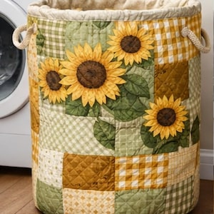 以下が含まれることがあります： ひまわりのデザインが施されたキルトのランドリーバスケット。バスケットは円筒形で、ベージュの縁とロープのハンドルが付いています。パッチワークのデザインは、ひまわりと葉のアップリケが付いた黄色、緑、茶色の正方形が特徴です。