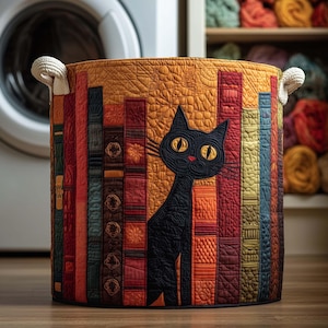 Op de afbeelding: Een ronde, gewatteerde opbergmand met een zwart kattenontwerp. De mand heeft een patchworkontwerp van kleurrijke boekruggen en een grote zwarte kat met gele ogen. De mand heeft witte touwhandvatten en wordt waarschijnlijk gebruikt voor wasgoed of opslag.