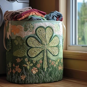 Può includere: Un cesto per la biancheria trapuntato verde e crema con un grande motivo a trifoglio. Il cesto è pieno di vestiti piegati di vari colori. Il design include trifogli e fiori ricamati. Il cesto ha due manici e si trova vicino a una finestra.
