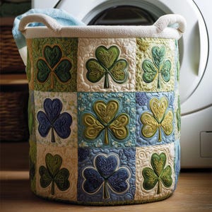 Può includere: Un cesto portabiancheria cilindrico con un design a trifoglio trapuntato. Il cesto presenta un bordo e manici color crema, e il corpo è composto da quadrati con motivi a trifoglio verdi, blu e crema. Un asciugamano blu è visibile all'interno.
