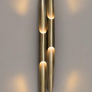 Peut inclure: Une applique murale verticale composée de deux tubes cylindriques allongés en laiton avec une finition métallique. Chaque tube présente trois découpes semi-circulaires qui émettent une lueur chaude et douce, créant un éclairage focalisé.