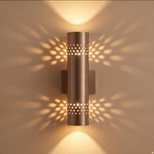 Op de afbeelding: Een cilindrische wandlamp met een metallic bronzen afwerking, voorzien van twee banden met geperforeerde ronde uitsparingen die een stervormig lichtpatroon op de muur boven en onder het armatuur werpen voor een warme, gerichte verlichting.