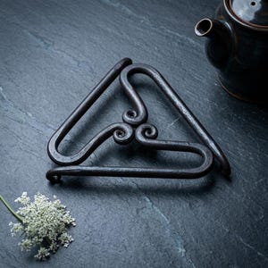 Könnte beinhalten: Ein dunkler, handgeschmiedeter Metalluntersetzer mit einem dreieckigen Rahmen und drei miteinander verbundenen, nach innen geschwungenen Spiralen in der Mitte. Die Metalloberfläche ist texturiert und matt, bestehend aus drei durchgehenden, geschlungenen Stäben.