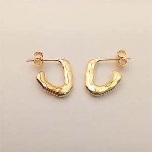 May include: A pair of gold-colored, square-shaped hoop earrings. Each earring has a post for piercing and a curved, textured design. The earrings are displayed on a light-colored surface.