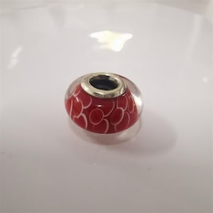 May include: A red and white glass bead with a silver-coloured metal rim. The bead features a pattern of white ovals on a red background. The bead is clear and transparent.