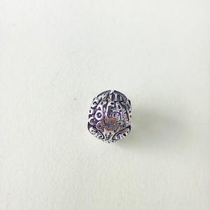 May include: A silver-coloured bead with an oval shape. The bead is covered in intricate, engraved patterns and text. The text appears to be a series of words or phrases. The bead is set against a white background.