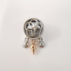 Puede incluir: Un charm de plata con un diseño circular que presenta una estrella de mar y formas entrelazadas. Tres colgantes de concha cuelgan debajo, con una concha de color dorado en el centro. El charm está sobre un fondo blanco.