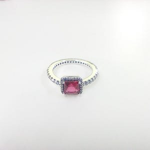 May include: A silver ring with a square, red gemstone center. The ring is surrounded by smaller, clear stones and has a delicate band. The ring is set against a white background, highlighting its design and details.