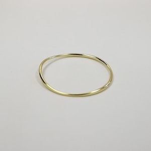 May include: A simple, gold-colored bangle bracelet. The bracelet has a smooth, slightly irregular surface and a circular shape. The jewelry is set against a plain, light background.