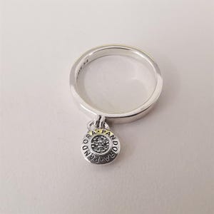 Pode incluir: Um anel de prata com um pequeno pingente redondo. O pingente tem uma parte superior dourada e uma base prateada com a palavra "PANDORA" ao redor da borda. O pingente é preenchido com pequenas pedras transparentes. O anel está em um fundo branco.