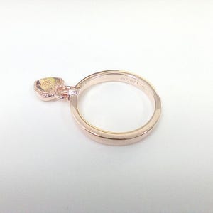 May include: Rose gold ring with a heart-shaped charm. The charm has a yellow center and a decorative border. The ring is set against a white background, showcasing its delicate design and color.