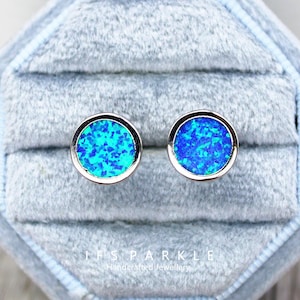 May include: A pair of round stud earrings with a silver-colored border. The center of each earring features a vibrant blue opal design. The earrings are displayed in a gray velvet jewelry box. The text "IFS SPARKLE Handcrafted Jewellery" is visible.