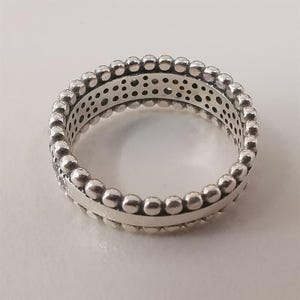 May include: A silver ring with a beaded design around the edges. The ring has a band of small, evenly spaced beads and a central section with a series of small holes. The ring is shown on a white surface.