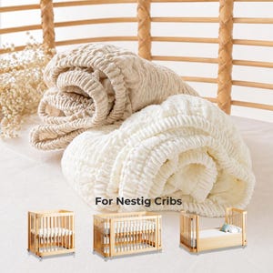 May include: Rolled beige and white crib sheets. The sheets are made of a textured fabric. The image also shows three small wooden cribs. Text on the image reads "For Nestig Cribs."