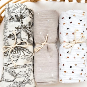 May include: Three rolled muslin blankets tied with cream-coloured rope. The left blanket features a black and white tropical leaf print. The centre blanket is solid beige with small white star patterns. The right blanket is white with tiny brown and black flower prints.