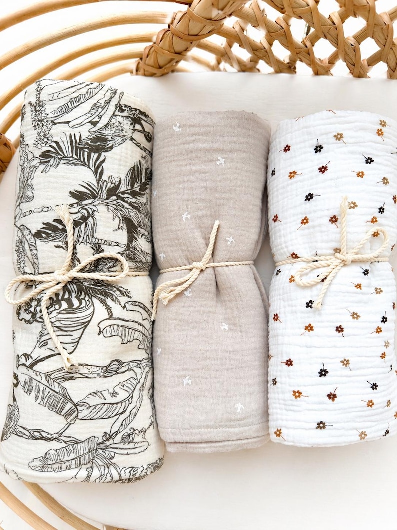 May include: Three rolled muslin blankets tied with cream-coloured rope. The left blanket features a black and white tropical leaf print. The centre blanket is solid beige with small white star patterns. The right blanket is white with tiny brown and black flower prints.