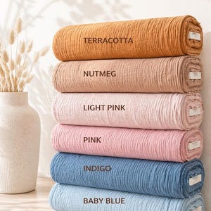 May include: A stack of six fabric rolls in various colours, each labelled with text: Terracotta, Nutmeg, Light Pink, Pink, Indigo, and Baby Blue. The fabric has a crinkled, textured surface and is rolled into cylindrical shapes with visible white cores.