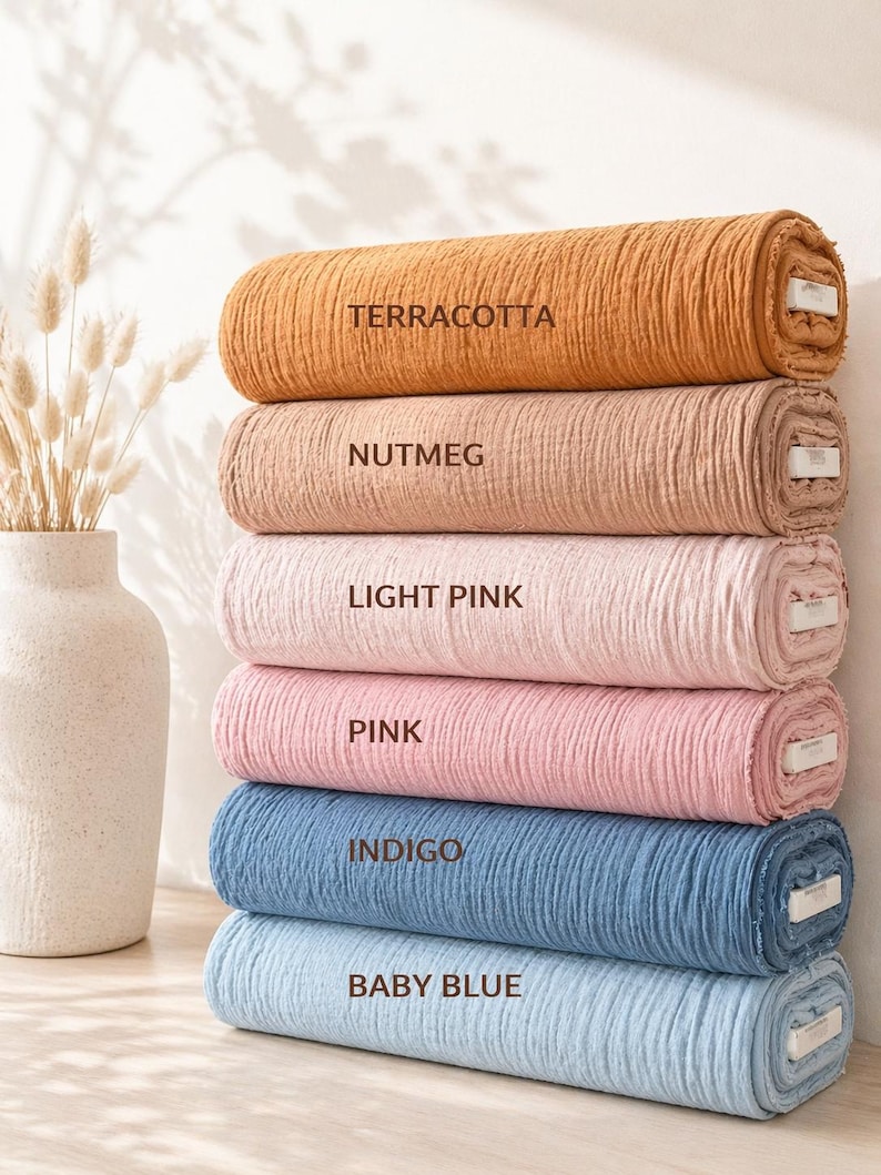 May include: A stack of six fabric rolls in various colours, each labelled with text: Terracotta, Nutmeg, Light Pink, Pink, Indigo, and Baby Blue. The fabric has a crinkled, textured surface and is rolled into cylindrical shapes with visible white cores.