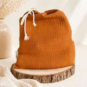 Könnte beinhalten: Ein orange-brauner Musselin-Kordelzugbeutel mit strukturierter Oberfläche. Der Beutel liegt auf einer runden Holzscheibe. Der Kordelzug ist weiß.