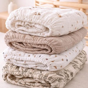 Puede incluir: Una pila de cuatro sábanas bajeras en varios colores neutros. La sábana superior es blanca con pequeños estampados florales marrones. La segunda sábana es de color beige liso. La tercera sábana es blanca con pequeños estampados florales morados y marrones. La sábana inferior tiene un estampado botánico.