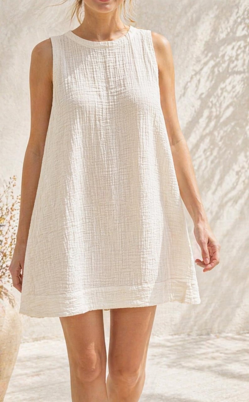 May include: A sleeveless, off-white mini dress featuring a textured, crinkled fabric finish. The garment has a high, rounded neckline and a straight-cut hemline, with a simple, loose-fitting silhouette that falls above the knee.