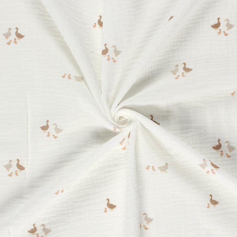 May include: A white muslin fabric featuring a repeating pattern of small brown and light grey geese and goslings. The textile has a crinkled, textured surface with a soft, gathered appearance radiating from the centre of the folded material.