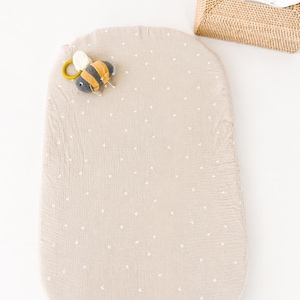May include: An oval-shaped beige changing pad cover with a small white cross pattern, featuring a plush bee-shaped toy with a yellow ring attached to the top left corner, positioned near a rectangular woven wicker tissue box holder.