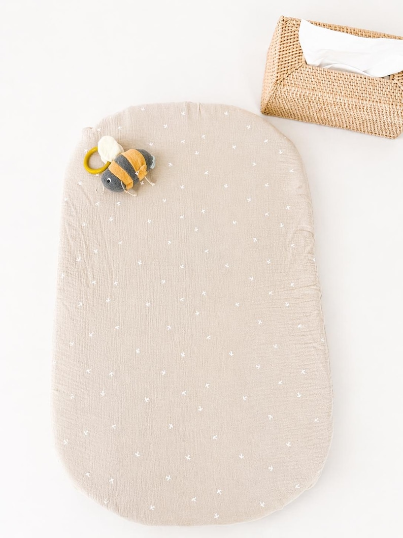 May include: An oval-shaped beige changing pad cover with a small white cross pattern, featuring a plush bee-shaped toy with a yellow ring attached to the top left corner, positioned near a rectangular woven wicker tissue box holder.