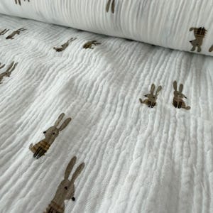 May include: A white crinkled muslin fabric featuring a repeating pattern of small, light brown rabbits. The textile has a soft, textured appearance with vertical folds and a lightweight, semi-sheer quality, showcasing the bunnies in various poses.