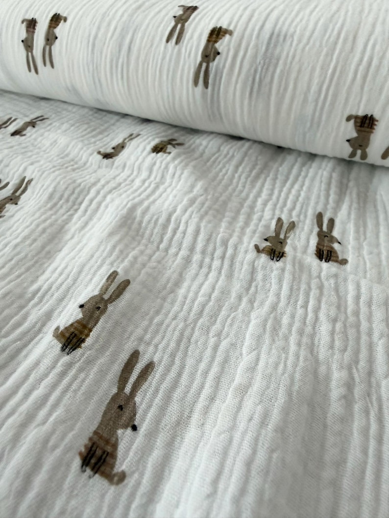 May include: A white crinkled muslin fabric featuring a repeating pattern of small, light brown rabbits. The textile has a soft, textured appearance with vertical folds and a lightweight, semi-sheer quality, showcasing the bunnies in various poses.