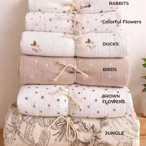May include: A stack of six folded muslin blankets tied with twine. The patterns include rabbits, colourful flowers, ducks, birds, brown flowers, and a jungle print. Each blanket is labelled with its respective design name in black text on the right side.