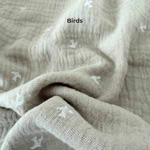 May include: A beige muslin fabric featuring a repeating pattern of small white embroidered birds. The textile has a crinkled, textured surface with folds and drapes. The word Birds is printed in black text on the fabric surface.