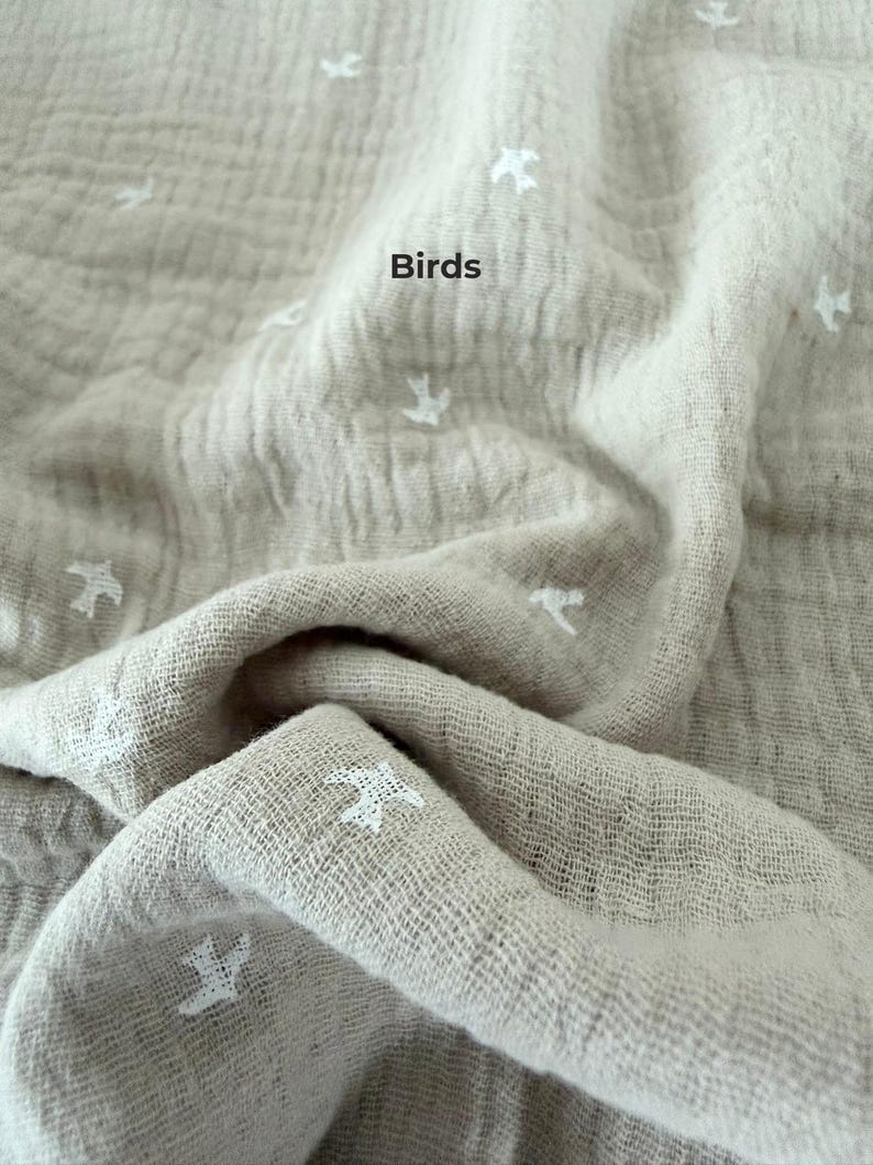 May include: A beige muslin fabric featuring a repeating pattern of small white embroidered birds. The textile has a crinkled, textured surface with folds and drapes. The word Birds is printed in black text on the fabric surface.