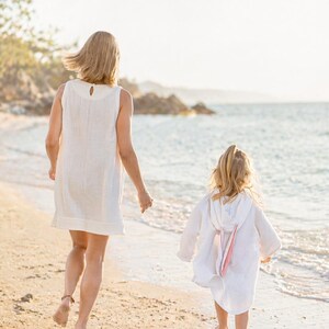 May include: A sleeveless white dress with a round neckline and a back button closure, and a white hooded cover-up featuring long sleeves and a soft pink ribbon detail on the back, both made of a lightweight, textured fabric worn on a sandy beach.