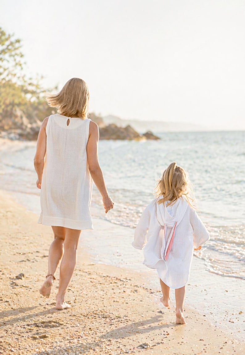 May include: A sleeveless white dress with a round neckline and a back button closure, and a white hooded cover-up featuring long sleeves and a soft pink ribbon detail on the back, both made of a lightweight, textured fabric worn on a sandy beach.
