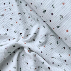 May include: A white double gauze fabric featuring a repeating pattern of small, scattered floral motifs in shades of muted orange, dark green, and brown. The textile has a distinct crinkled, puckered texture throughout the entire surface.