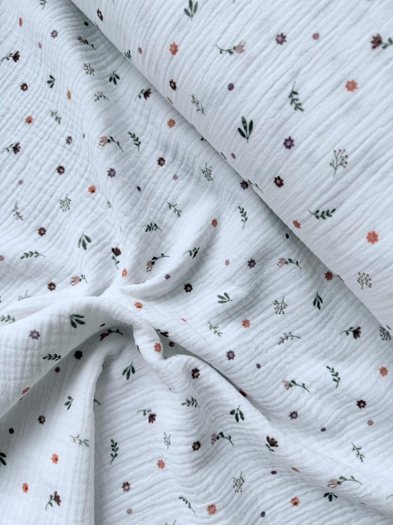 May include: A white double gauze fabric featuring a repeating pattern of small, scattered floral motifs in shades of muted orange, dark green, and brown. The textile has a distinct crinkled, puckered texture throughout the entire surface.