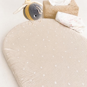 May include: A beige oval changing mat cover with a small white floral pattern, accompanied by a plush bee toy hanging from a yellow ring, a woven tissue box holder, and a folded white cloth with a subtle pink floral print on a white surface.