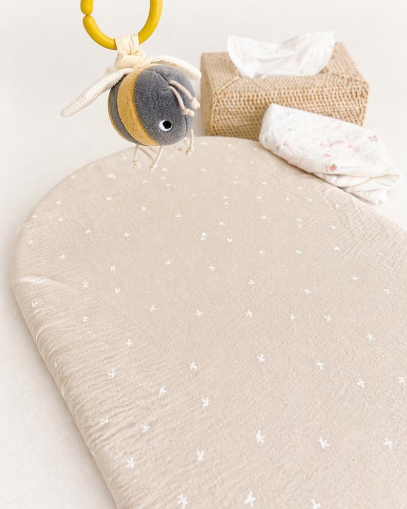 May include: A beige oval changing mat cover with a small white floral pattern, accompanied by a plush bee toy hanging from a yellow ring, a woven tissue box holder, and a folded white cloth with a subtle pink floral print on a white surface.