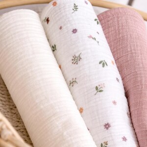 May include: Three rolled muslin swaddle blankets featuring a textured fabric. One blanket is solid cream, one is white with a small floral pattern in pink, orange, and green, and one is a solid dusty rose colour. The blankets are arranged side by side.