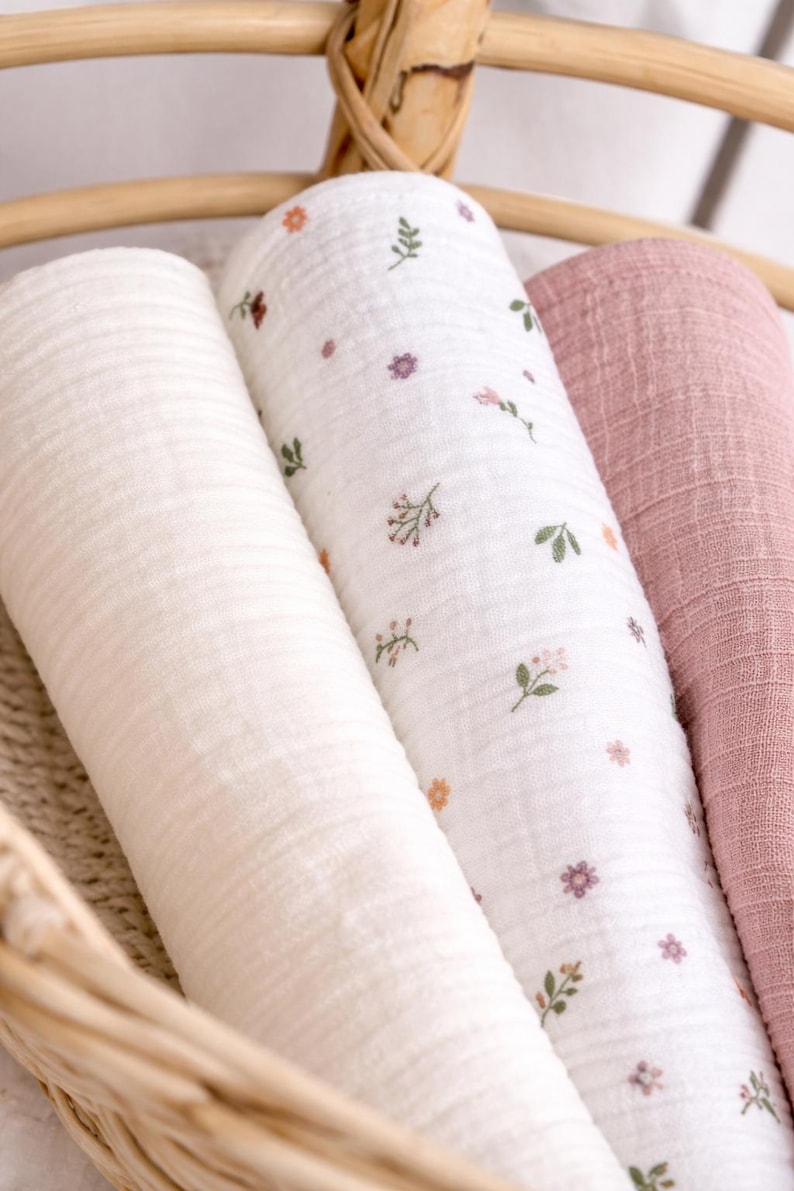 May include: Three rolled muslin swaddle blankets featuring a textured fabric. One blanket is solid cream, one is white with a small floral pattern in pink, orange, and green, and one is a solid dusty rose colour. The blankets are arranged side by side.