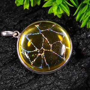 May include: A round pendant featuring a smooth, translucent yellow surface with an embedded iridescent, silver-toned web-like pattern. The circular piece is encased in a polished silver-coloured metal frame with a small loop at the top for attachment.