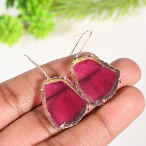 Puede incluir: Pendientes de plata con una vibrante piedra preciosa rosa. Las piedras preciosas tienen una forma irregular con un borde dorado. Los pendientes se presentan sobre un fondo neutro, resaltando su color y diseño.