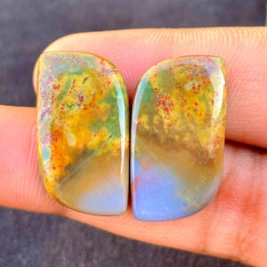 May include: Two polished, teardrop-shaped cabochons featuring a blend of colors including mustard yellow, moss green, and soft lavender. The stones display intricate, natural inclusions with small red speckles scattered across the surface.