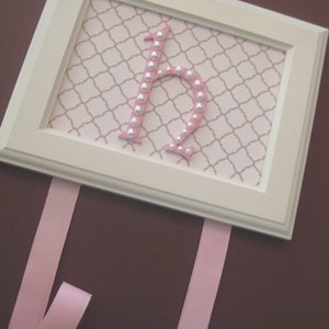 May include: A white frame with a pink ribbon on top. Inside the frame is a pink letter "H" made of pearl-like beads on a white background with a brown trellis pattern.