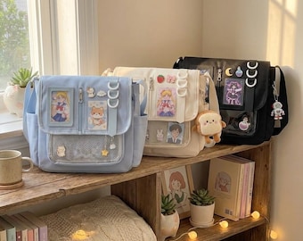 Könnte beinhalten: Drei Messenger-Taschen in Pastellfarben: Hellblau, Creme und Schwarz. Jede Tasche hat dekorative Elemente wie Charakterkarten, Charms und eine Netztasche. Die Taschen stehen auf einem Holzregal mit Büchern und Dekorationsartikeln.